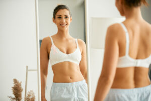 Woman in white bra with pink breast cancer ribbon looking in the mirror and considering immediate vs. delayed breast reconstruction