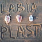 The word "labiaplasty" written in the sand, with three seashells positioned around it