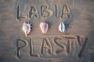 The word "labiaplasty" written in the sand, with three seashells positioned around it